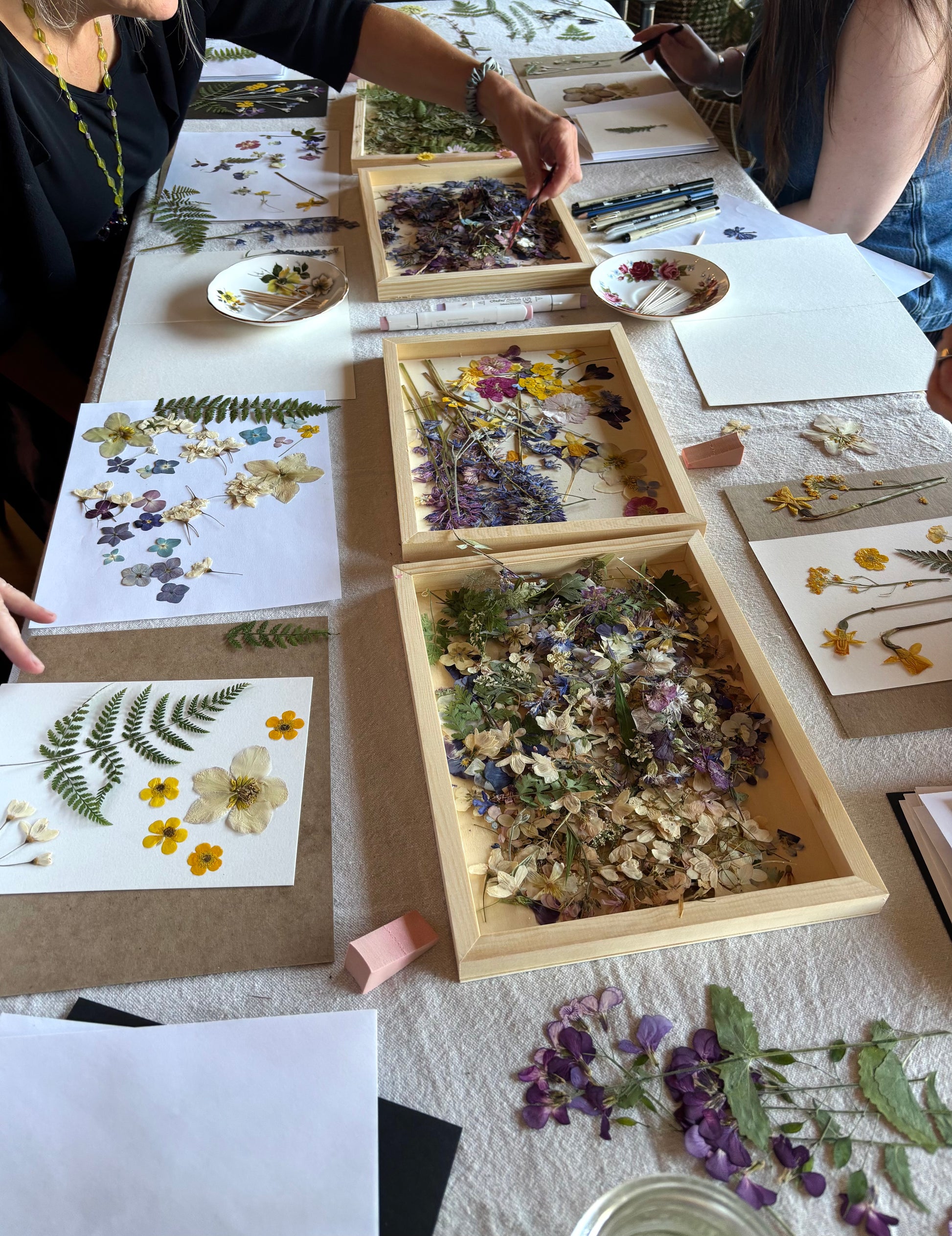 people creating pressed flower art from a variety of pressed flowers displayed in wooden trays
